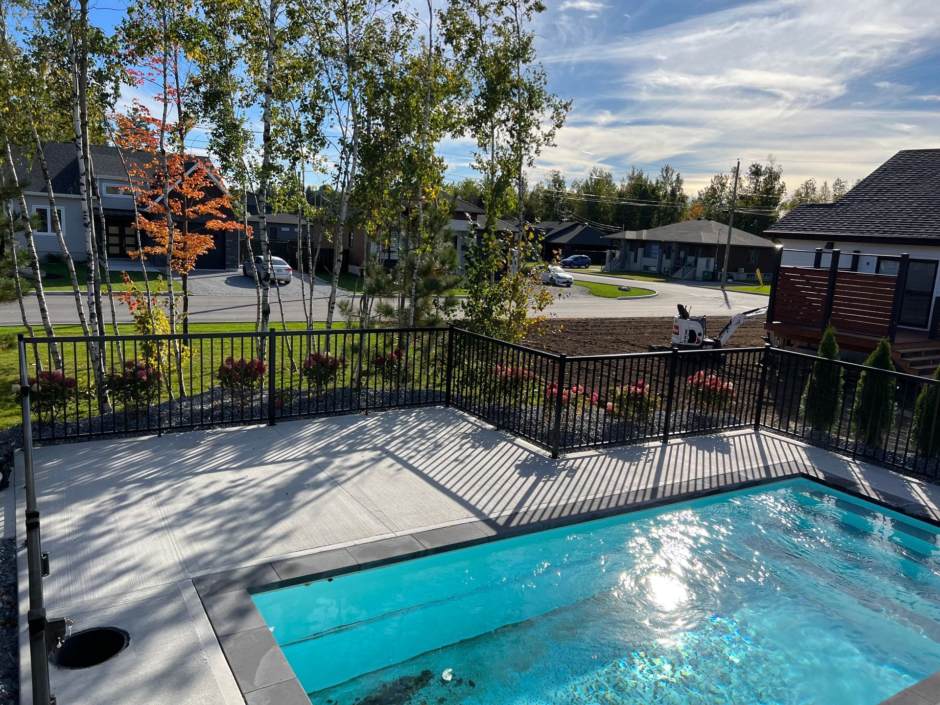 Piscine dans le jardin avec eau bleue, clôture noire, arbres et maisons voisines par une journée ensoleillée