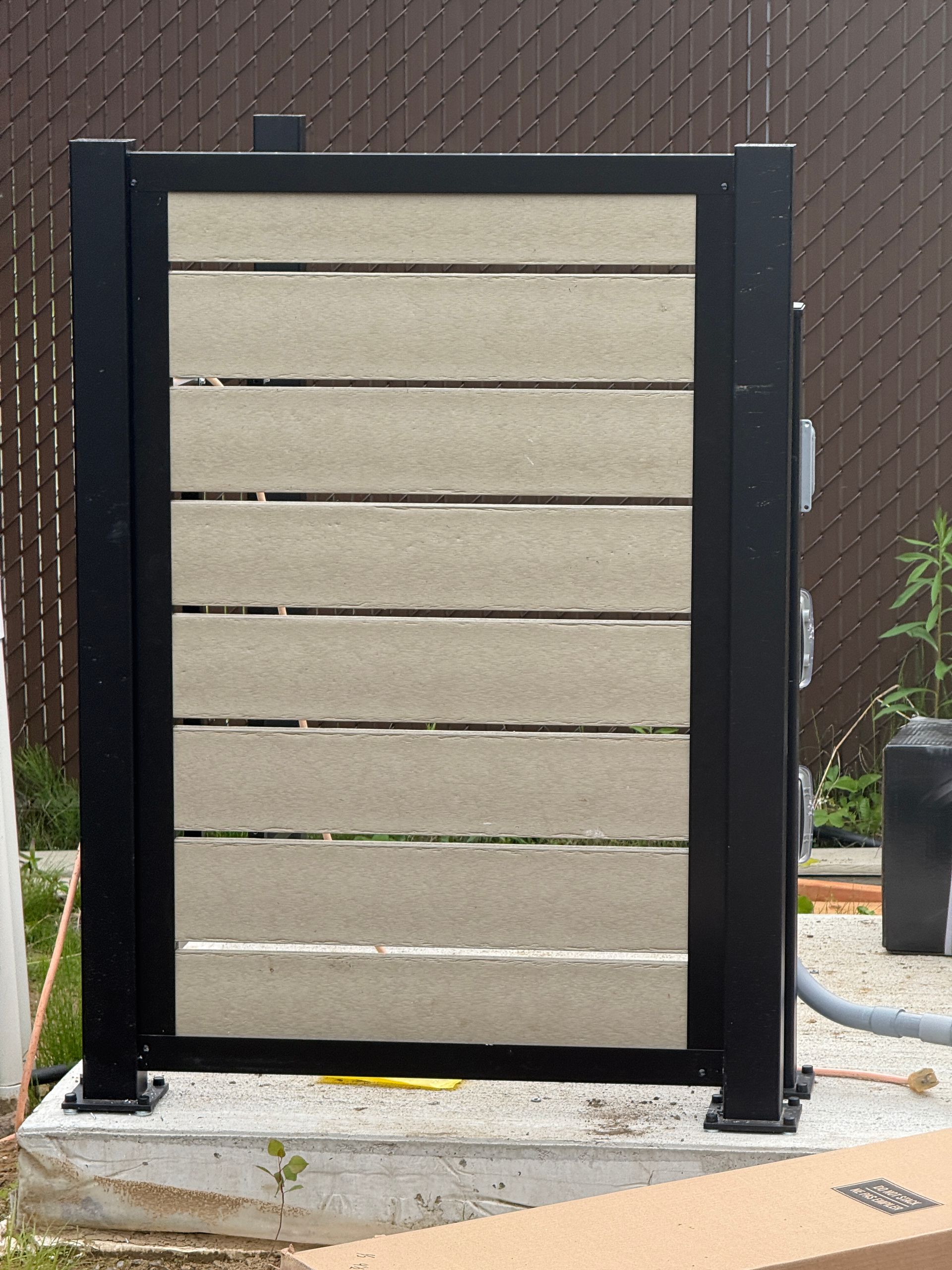 Panneau utilitaire extérieur noir et beige, fixé sur une base en béton, à côté d'un tuyau d'arrosage et d'un mur en briques.