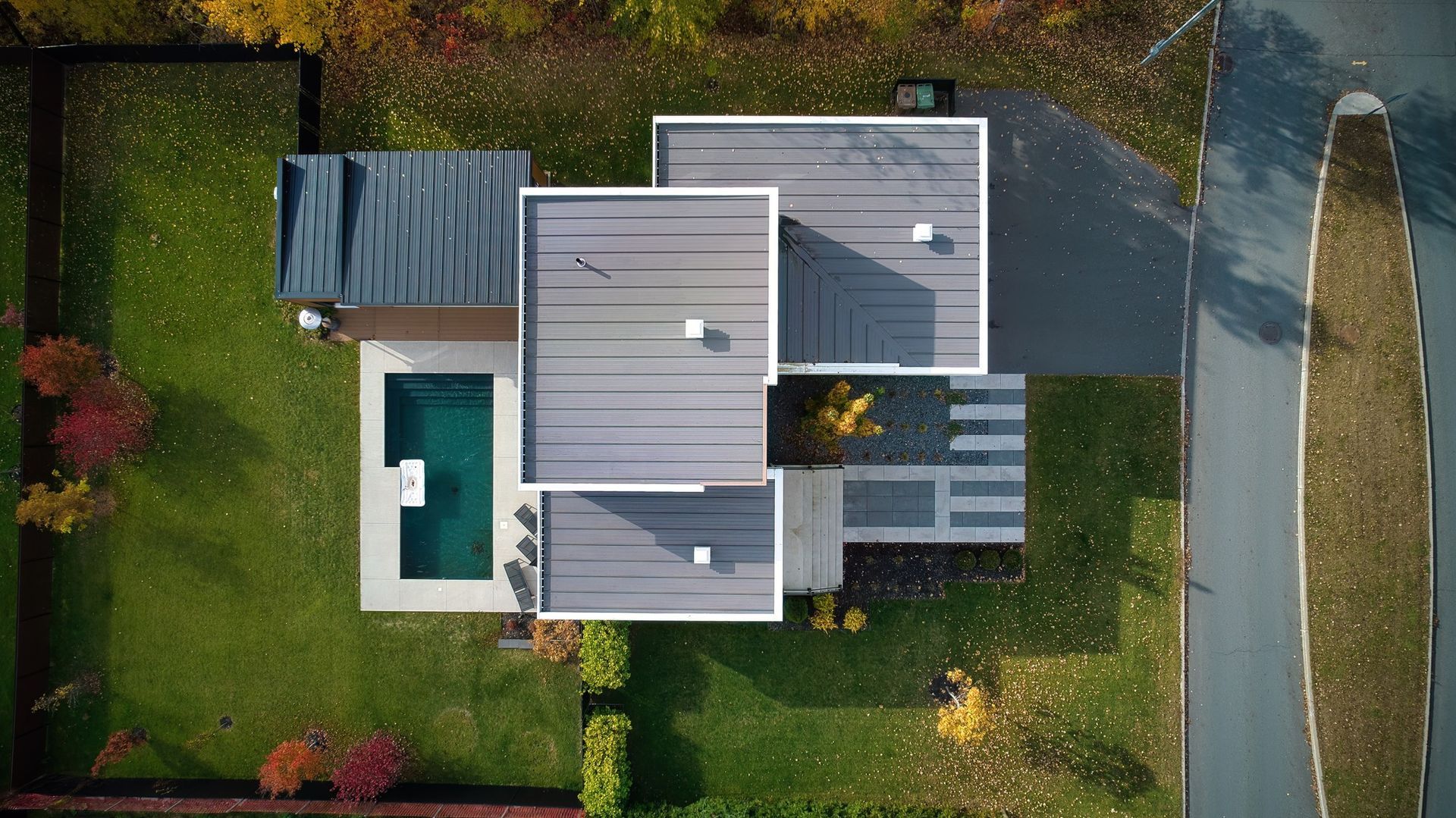 Vue aérienne d'une maison moderne avec une petite piscine, une terrasse et une pelouse environnante en bordure de route.