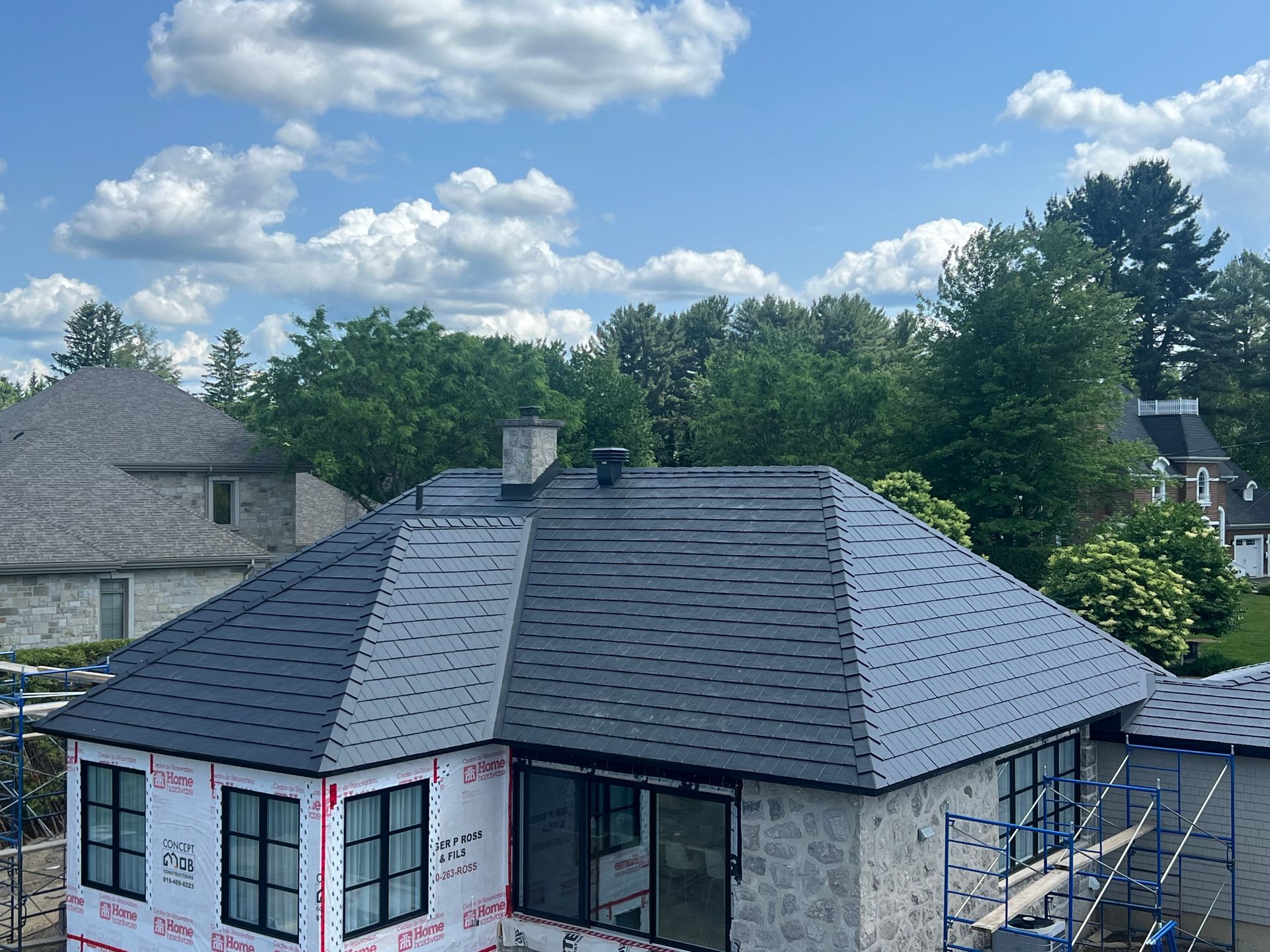 Maison partiellement construite avec un toit gris foncé et un revêtement rose, entourée d'arbres sous un ciel bleu nuageux