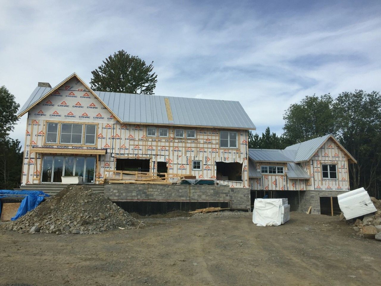 Maison partiellement construite avec un toit gris foncé et un revêtement rose, entourée d'arbres sous un ciel bleu nuageux