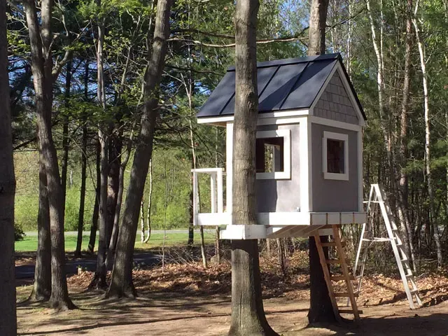 Petite cabane grise perchée dans un arbre, avec un toit noir, dans une zone boisée, à côté d'une échelle et d'arbres.
