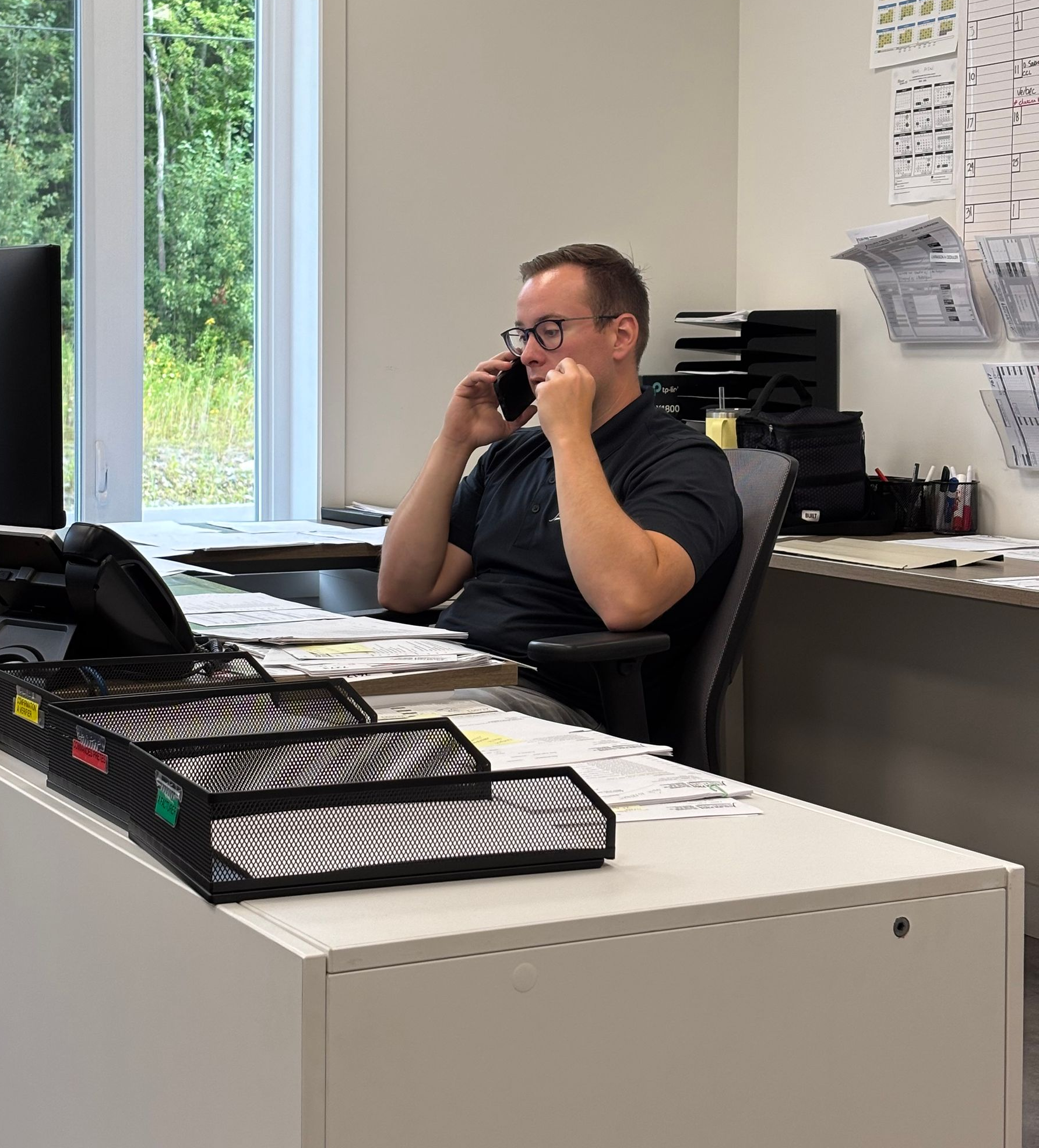 Un homme assis à un bureau parle au téléphone, avec des papiers et des plateaux de classement au premier plan.