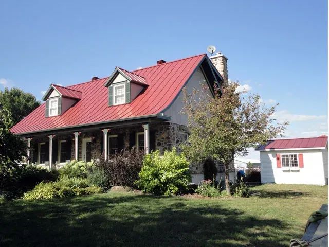 Maison en pierre au toit rouge et au porche, située dans une cour ensoleillée, avec une petite dépendance blanche à proximité.