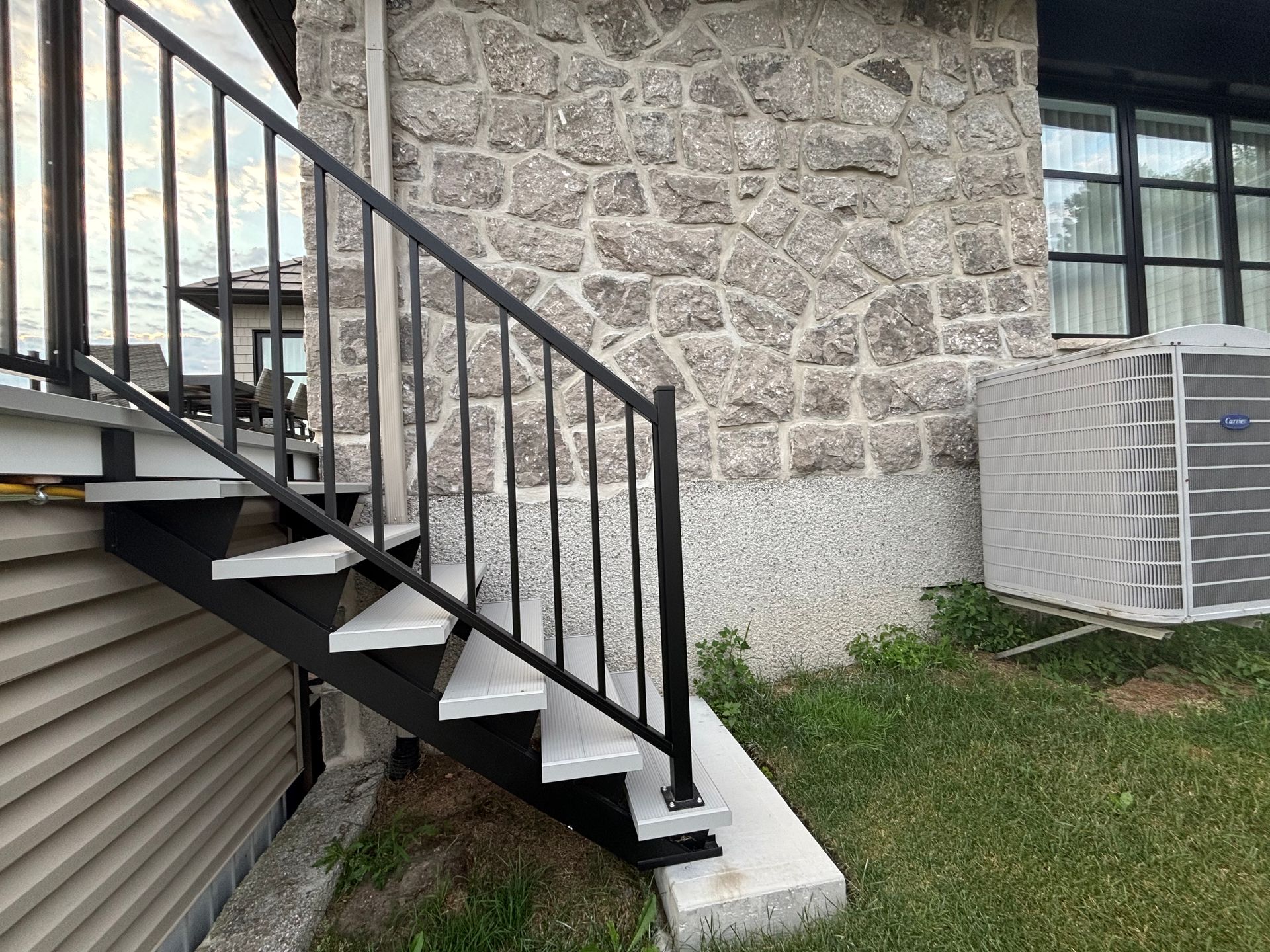 Escalier extérieur en métal, adossé à un mur en pierre et à une fenêtre, à côté d'une porte de garage et d'un climatiseur.