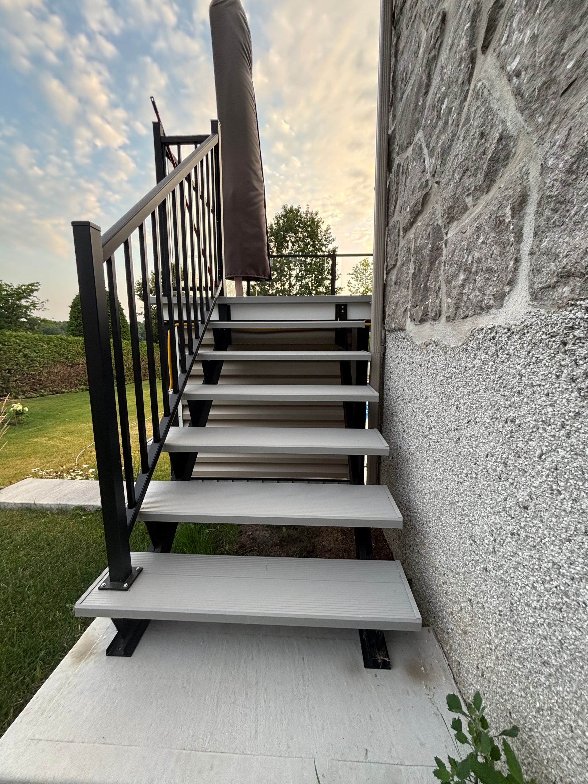 Escalier extérieur en métal, adossé à un mur en pierre, avec rampes noires et marches blanches.