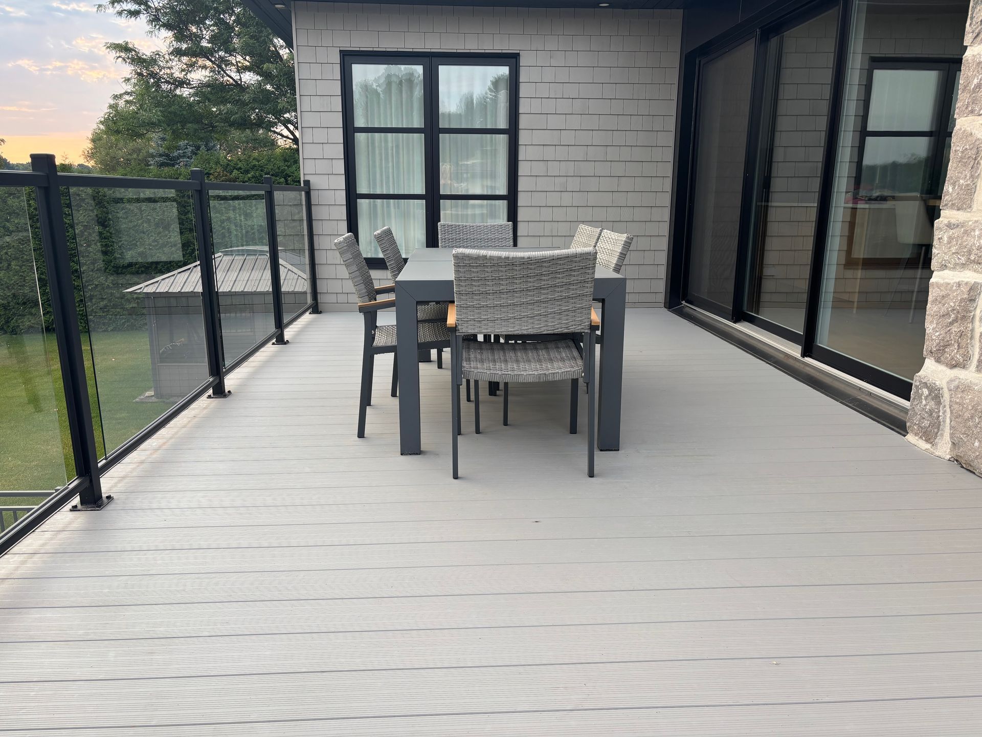 Terrasse extérieure avec chaises et table grises à côté d'une maison, donnant sur une cour clôturée au crépuscule