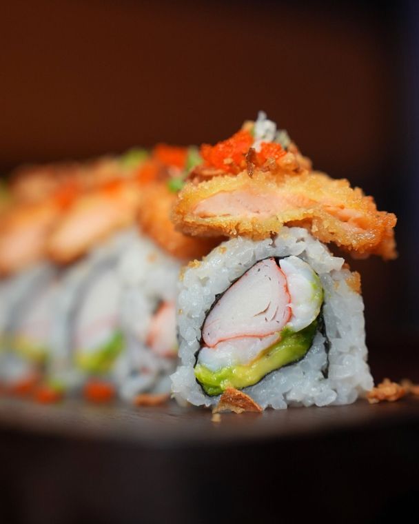 Close-up of sushi rolls topped with orange garnish on a dark plate, with blurred sushi in the background
