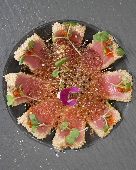 Overhead view of a round sushi platter with salmon slices, rice, herbs, and edible flowers on a dark plate