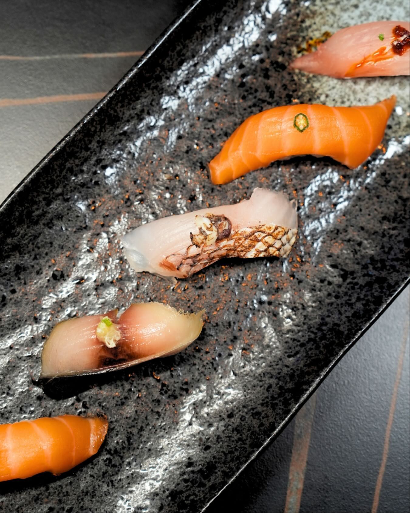 Assorted sushi pieces on a black rectangular plate, including salmon and seared fish.