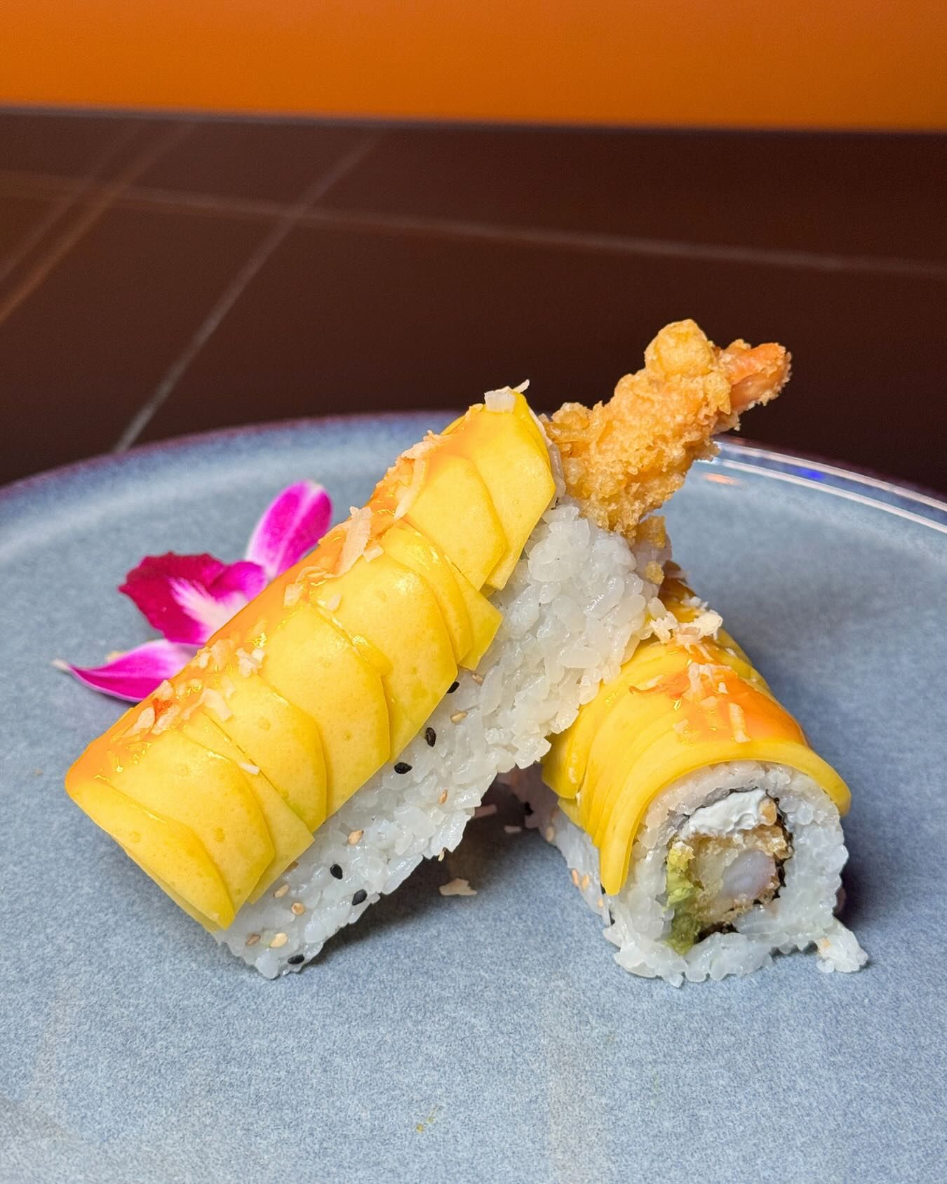 Sushi rolls with yellow mango topping on a gray plate, garnished with pink flowers