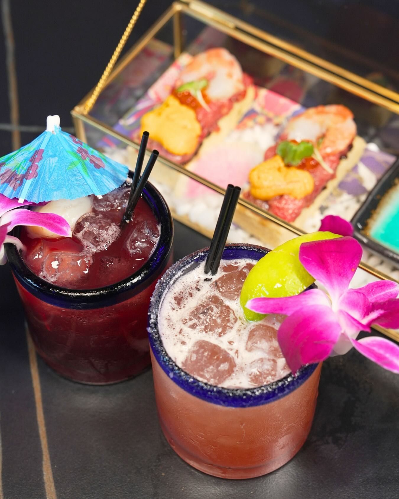 Two colorful cocktails with black straws, one garnished with a purple umbrella and one with an orchid, beside sushi