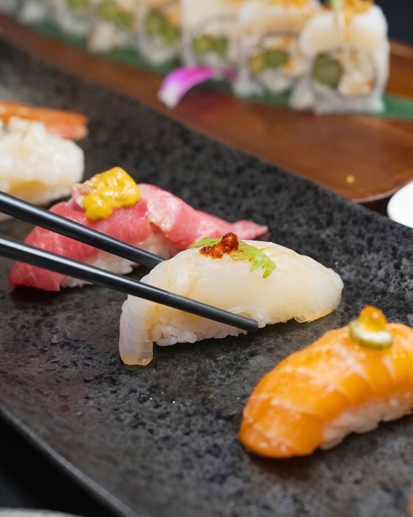 Chopsticks lifting nigiri sushi on a black plate, with colorful sushi pieces in the background