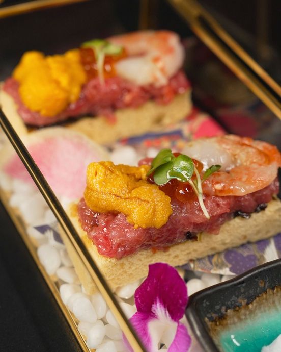 Two sushi-like bites topped with shrimp, sea urchin, and herbs on a dark tray