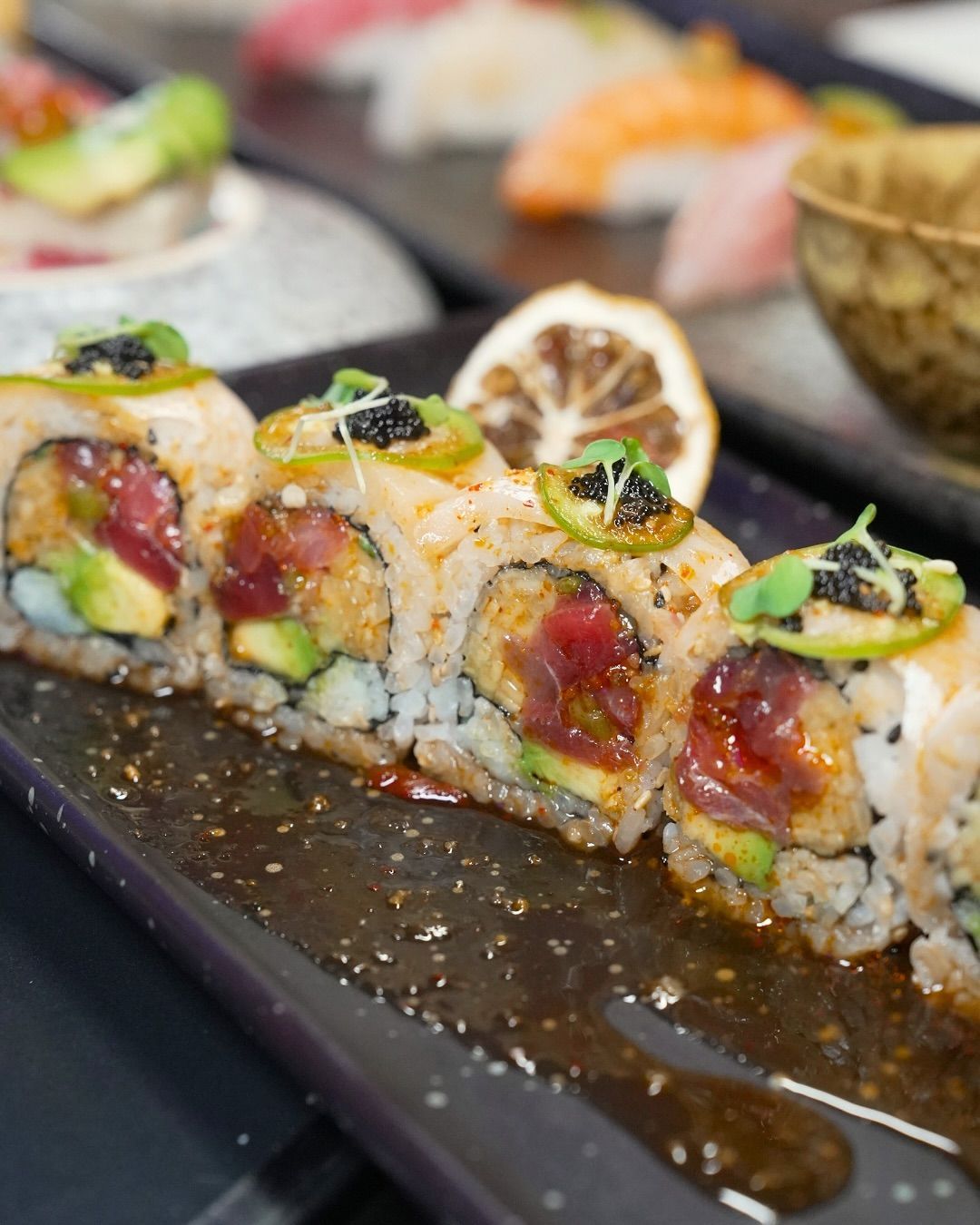 Sushi roll slices topped with jalapeño and lemon on a black plate, with dipping sauce and sushi in the background
