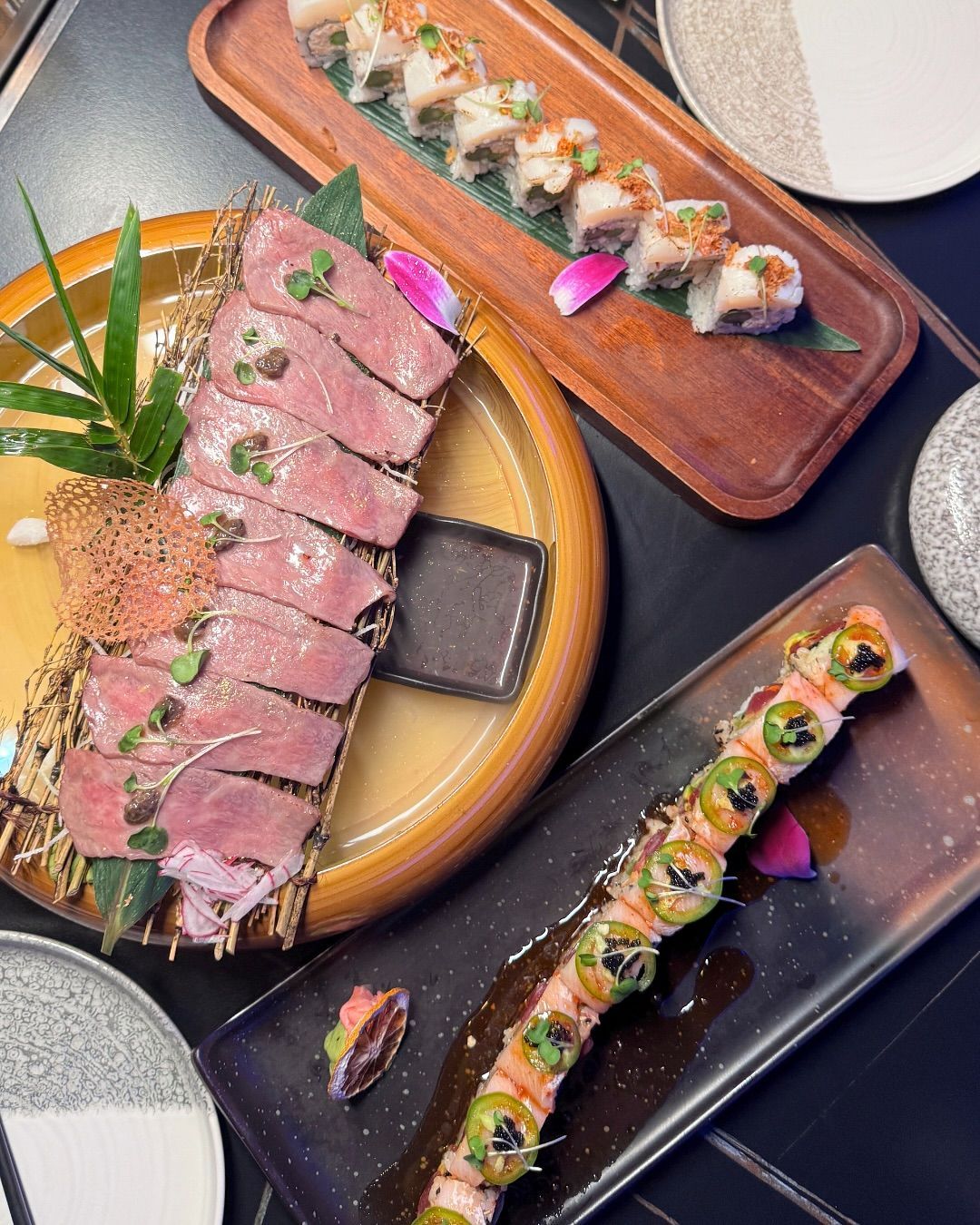Plated sushi and sliced steak appetizers on dark serving boards and a round plate.