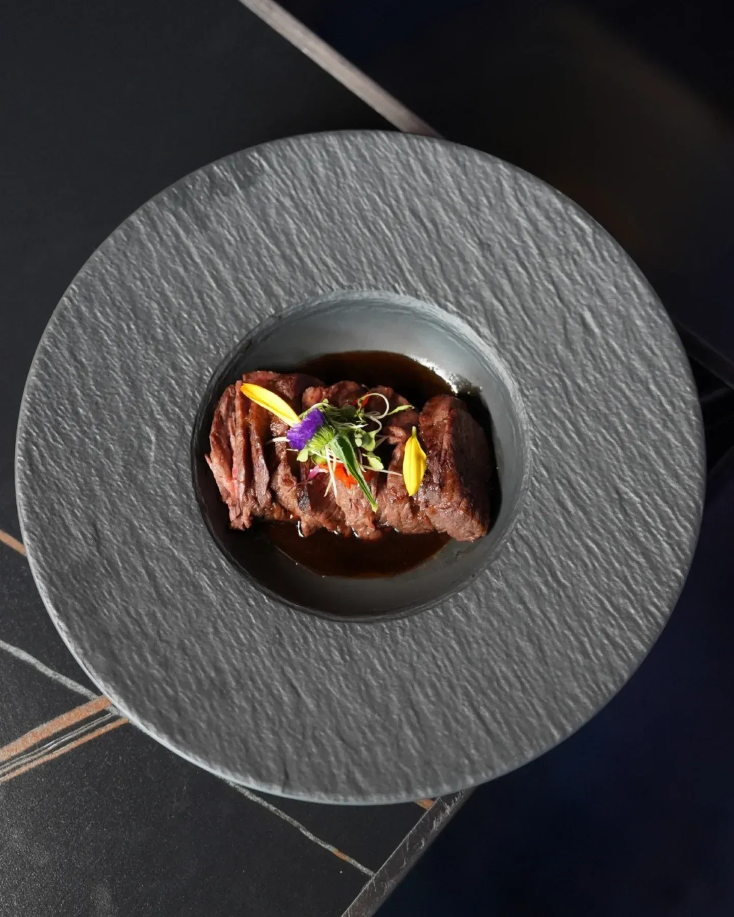Seared meat garnished with herbs on a black plate, viewed from above.