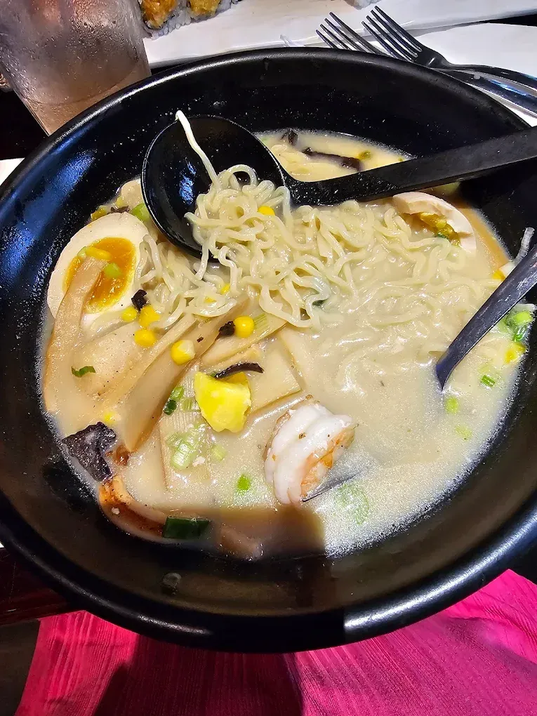 Black bowl of creamy noodle soup with seafood, mushrooms, corn, and chopsticks on a table