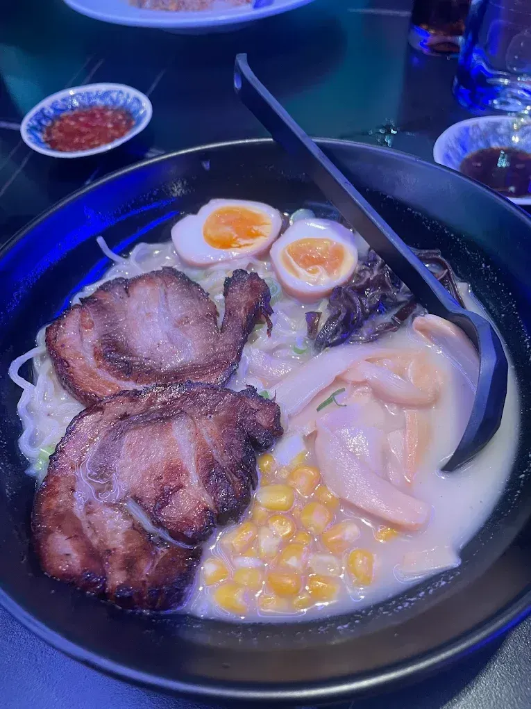 Ramen bowl with sliced pork, soft-boiled eggs, corn, and bamboo shoots, topped with chopsticks.
