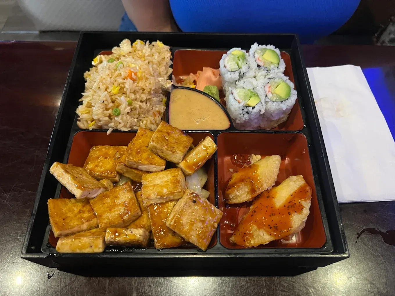 Bento box with sushi rolls, fried rice, tofu squares, and saucy chicken on a table