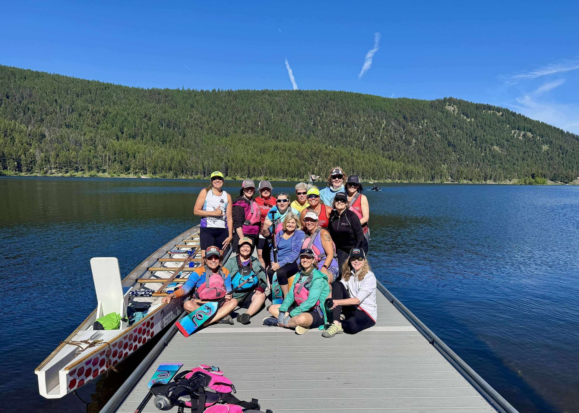 Group of people on a dock beside a dragon boat on a lake. Blue sky, mountains in background.