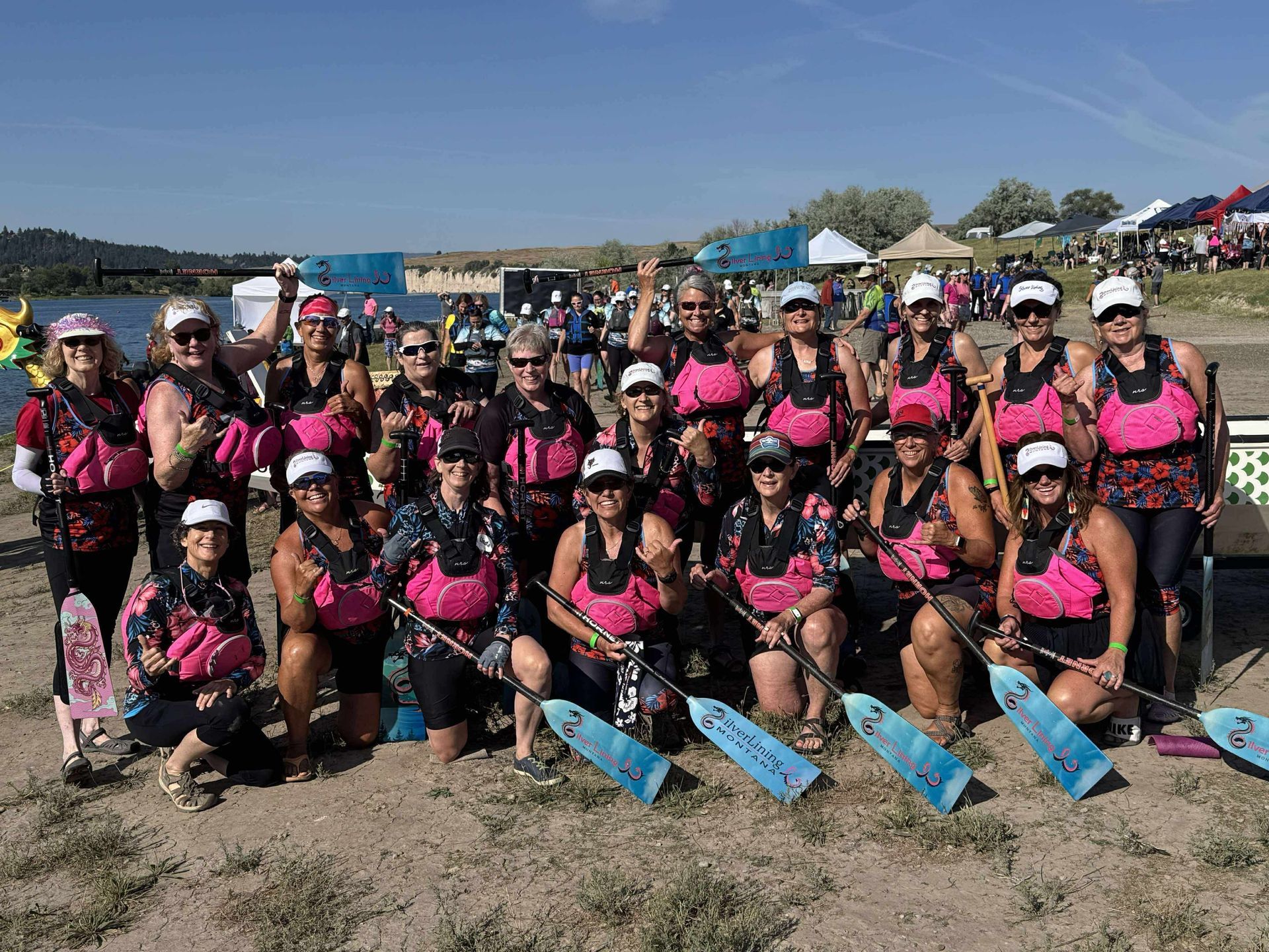 Dragon boat team in pink vests and hats, holding paddles near a lake.