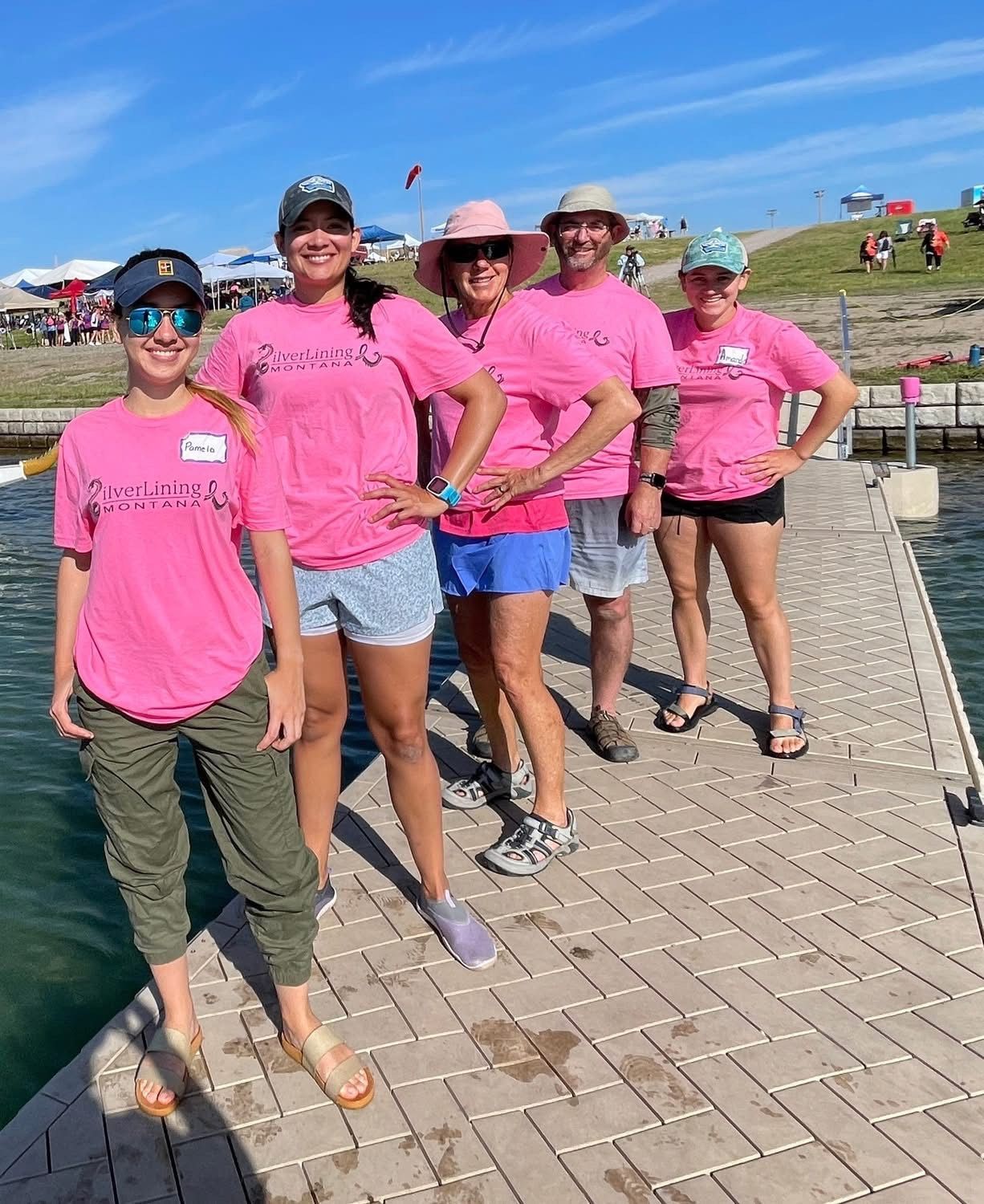 Five people in pink shirts stand on a dock. They smile. Sunny day at a lakeside event.