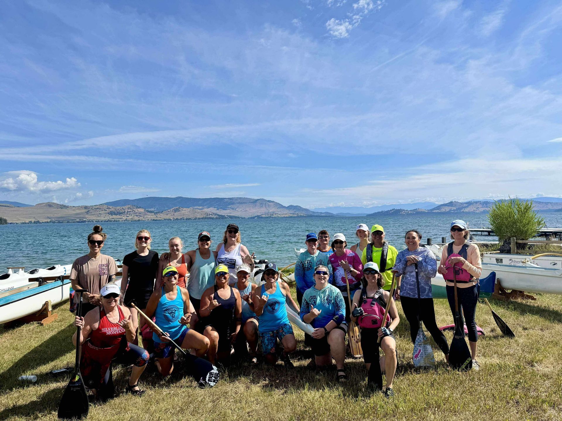Group of people pose on a grassy shore with boats, lake, and hills under a blue sky.