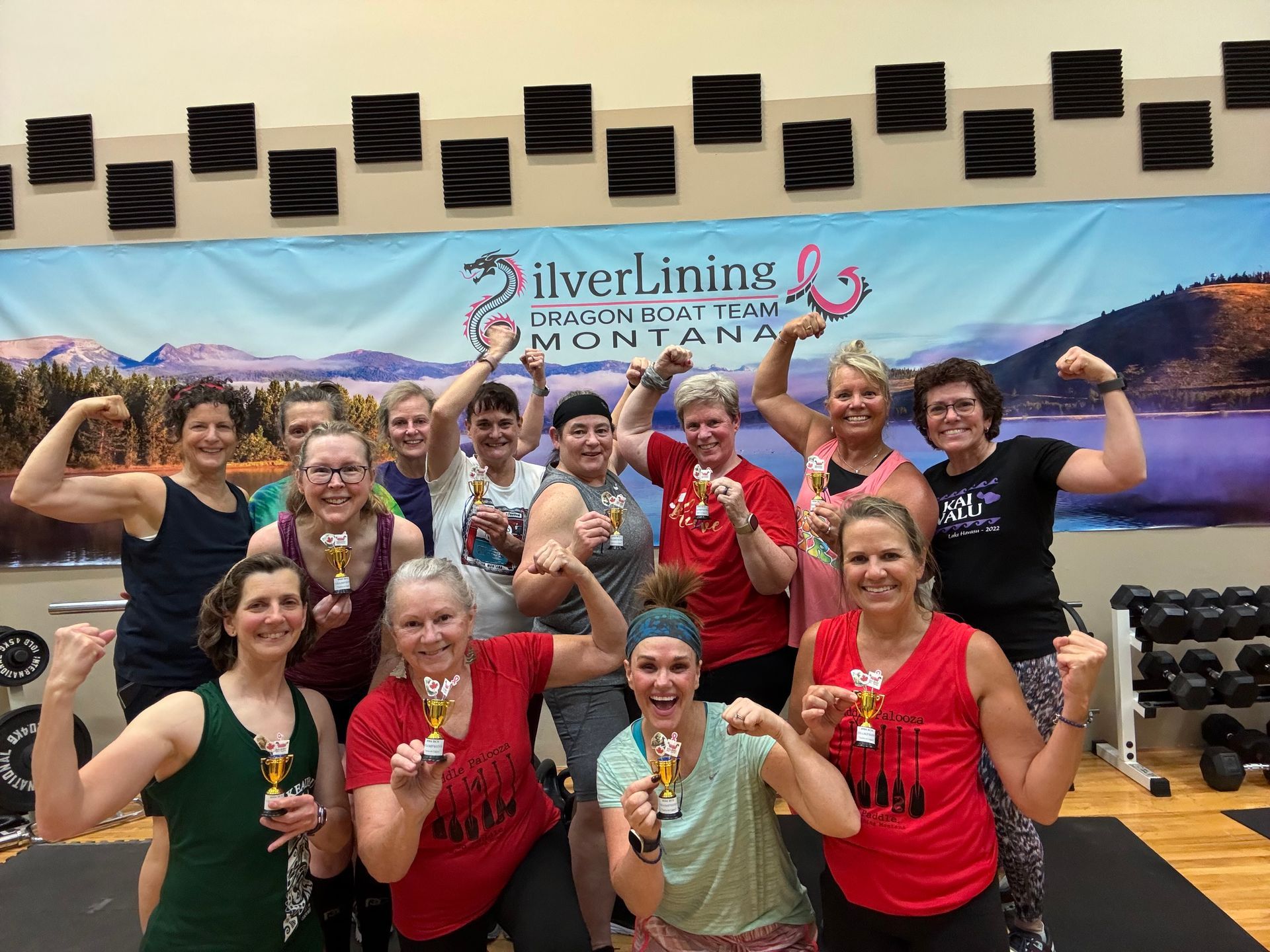 Group of women in gym flexing, holding trophies.