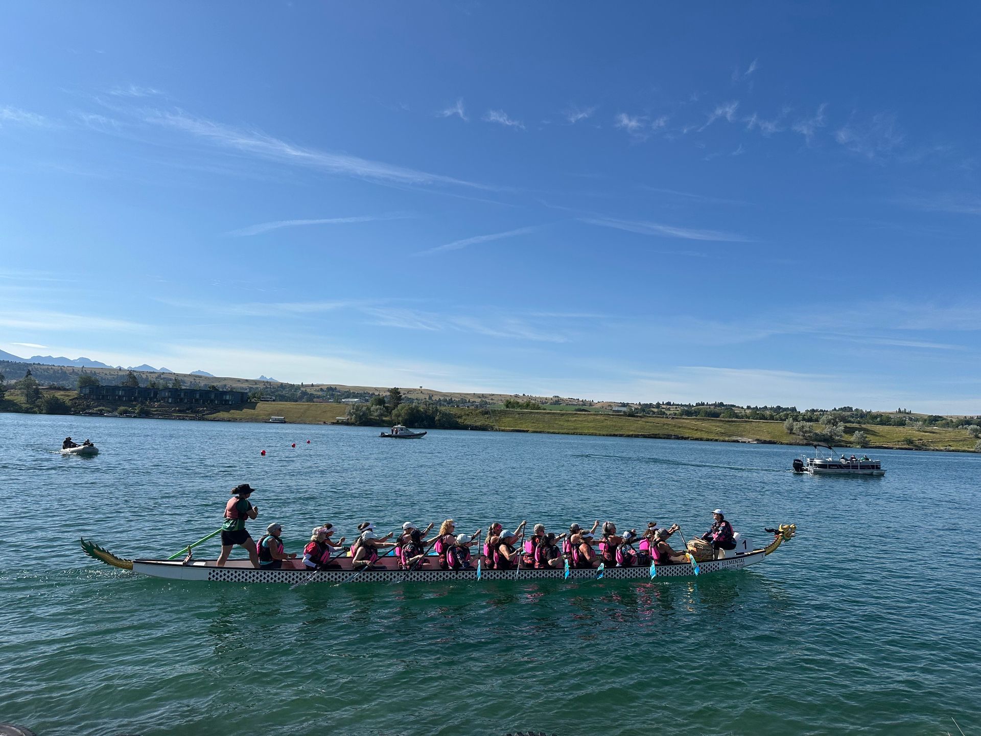 Dragon boat on water with paddlers, blue sky and shoreline.