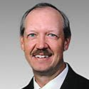 Headshot of a man with a mustache, wearing a white collared shirt and black blazer against a grey background.