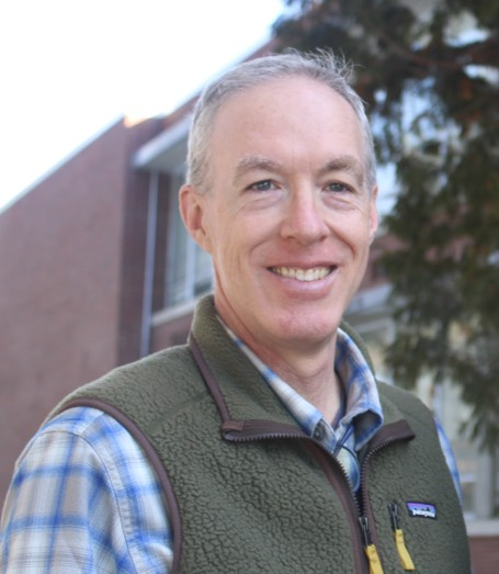 A smiling person with gray hair wearing a blue plaid shirt and a green fleece vest, standing outdoors near a building.