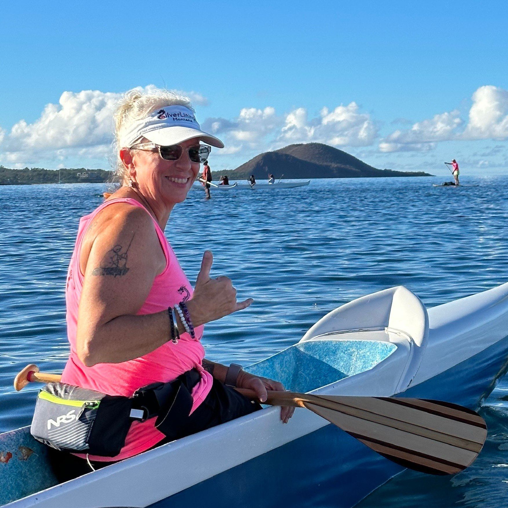 A person in a bright pink tank top and white visor sits in an outrigger canoe on the water, giving a shaka sign.