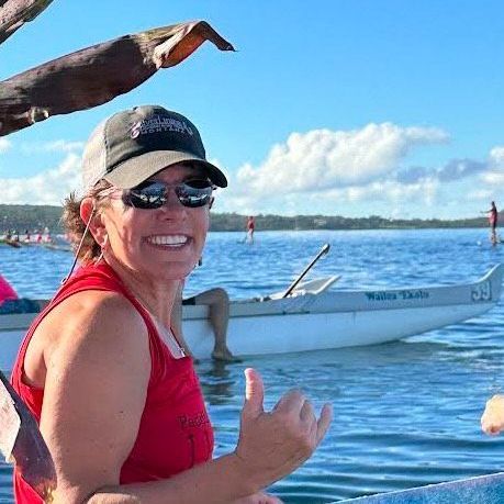 A smiling person in a red tank top and baseball cap makes a shaka sign in front of an outrigger canoe on a sunny bay.