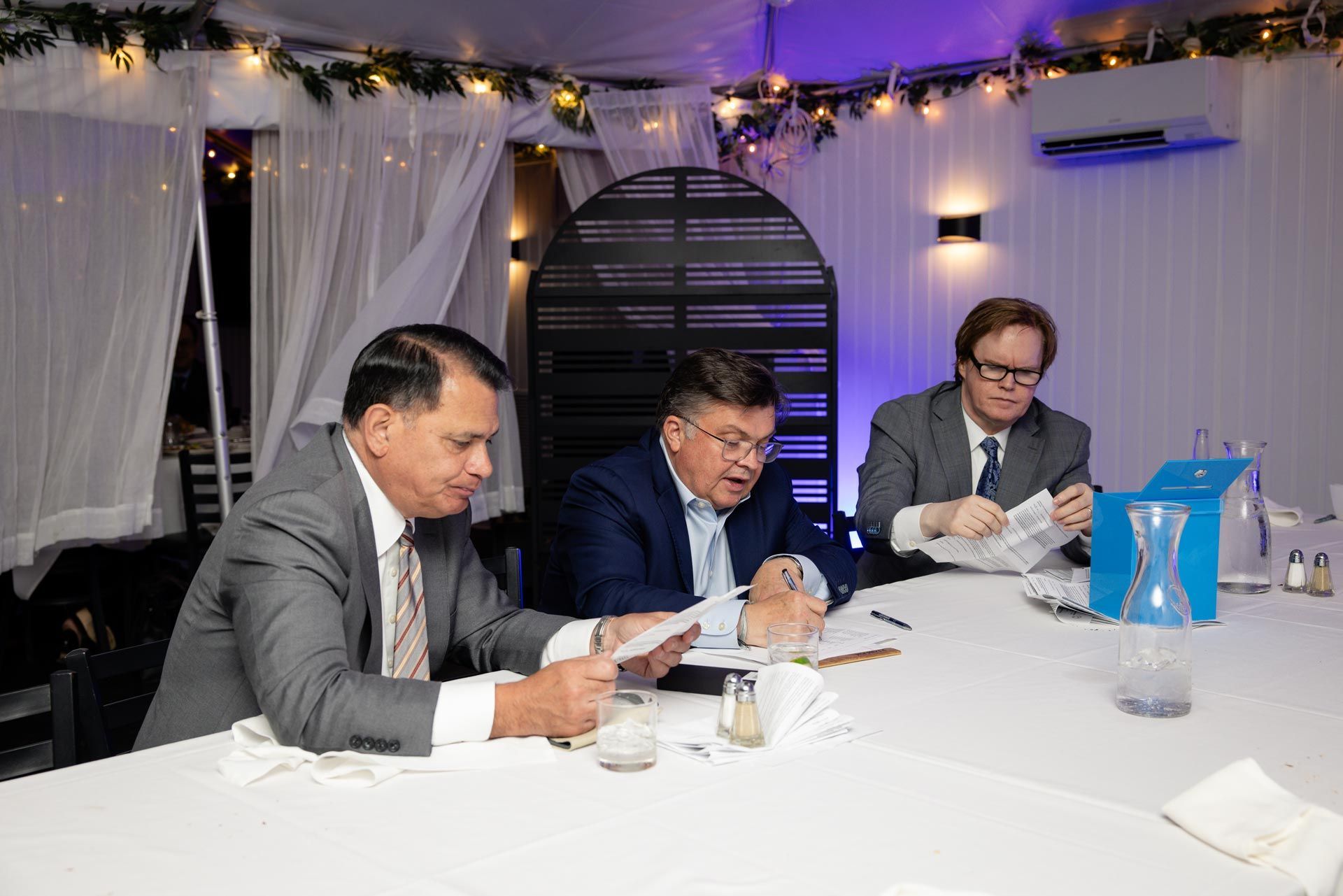 Three men seated at a table signing documents at an indoor event with white drapes and string lights