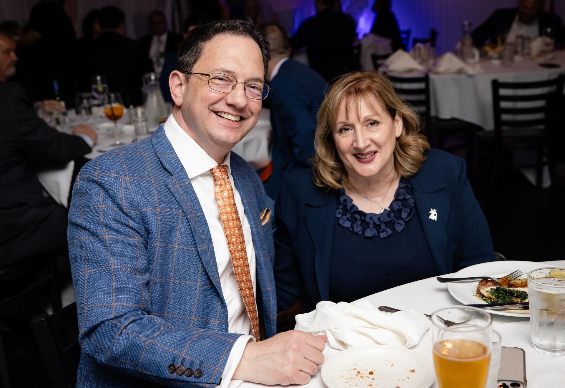 Two people smiling at a banquet table with drinks and plates in a dimly lit event hall