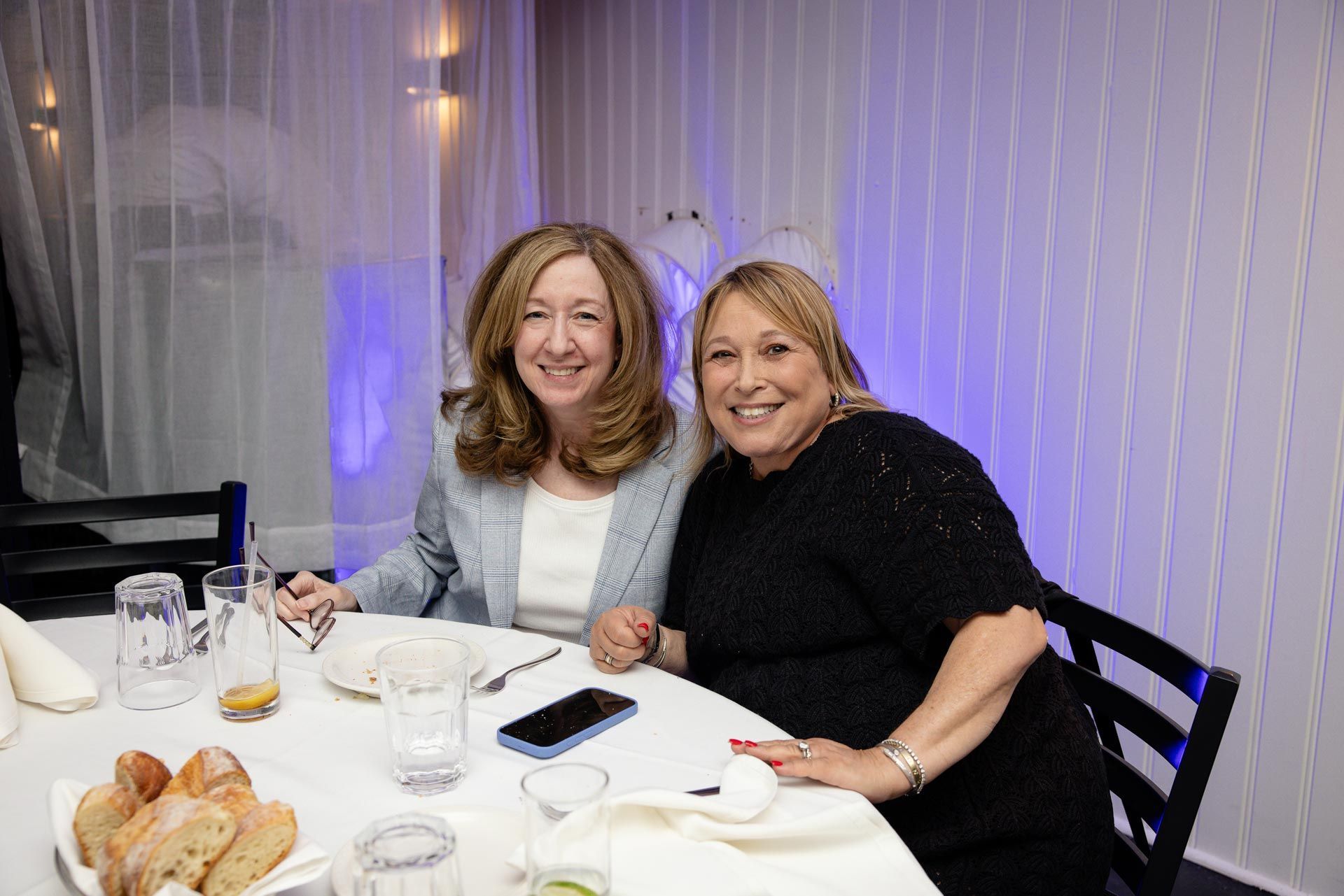 Two people smiling at a table in a softly lit restaurant, with plates, glasses, and a phone nearby