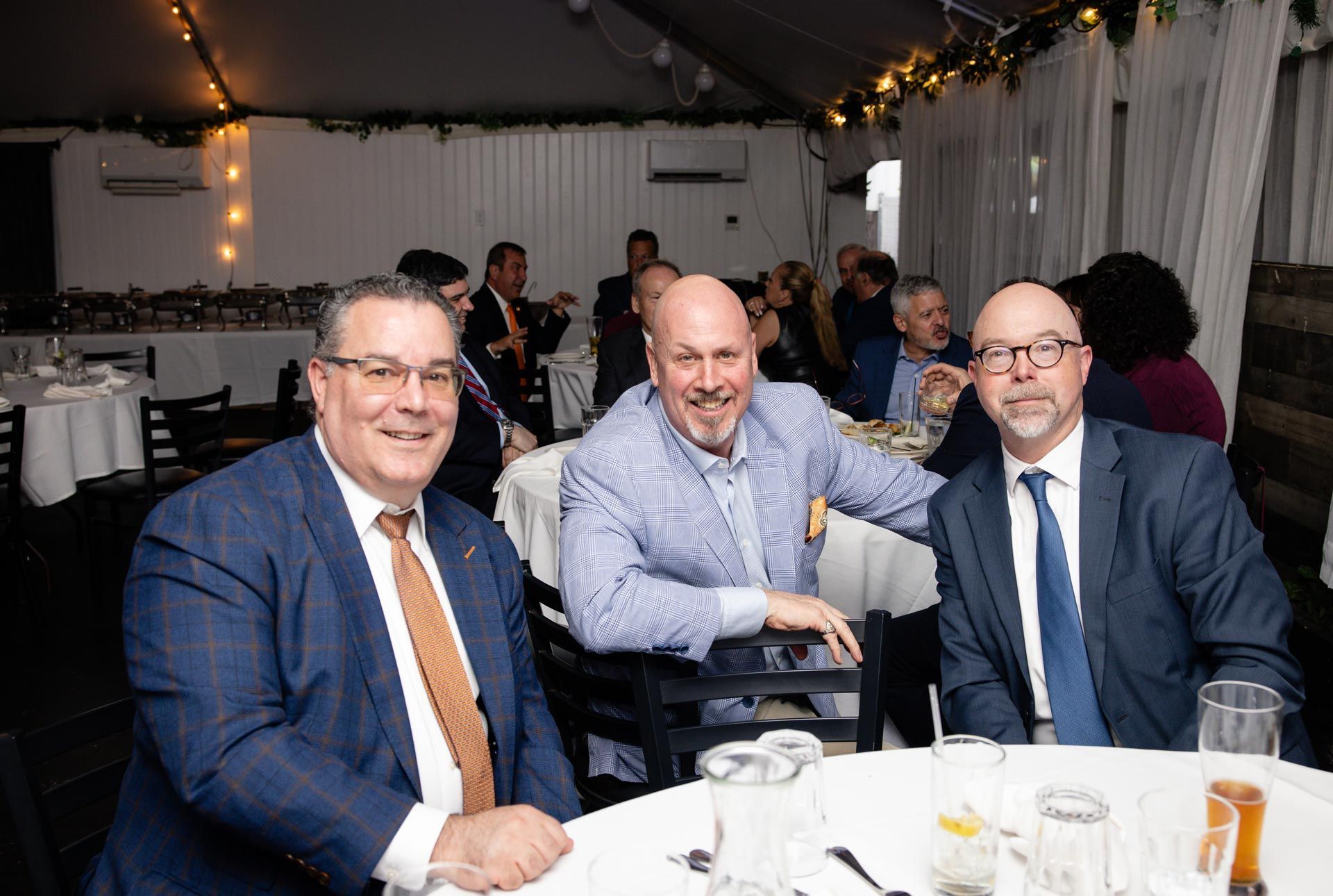 Three men seated at a banquet table under a decorated tent, smiling at the camera.
