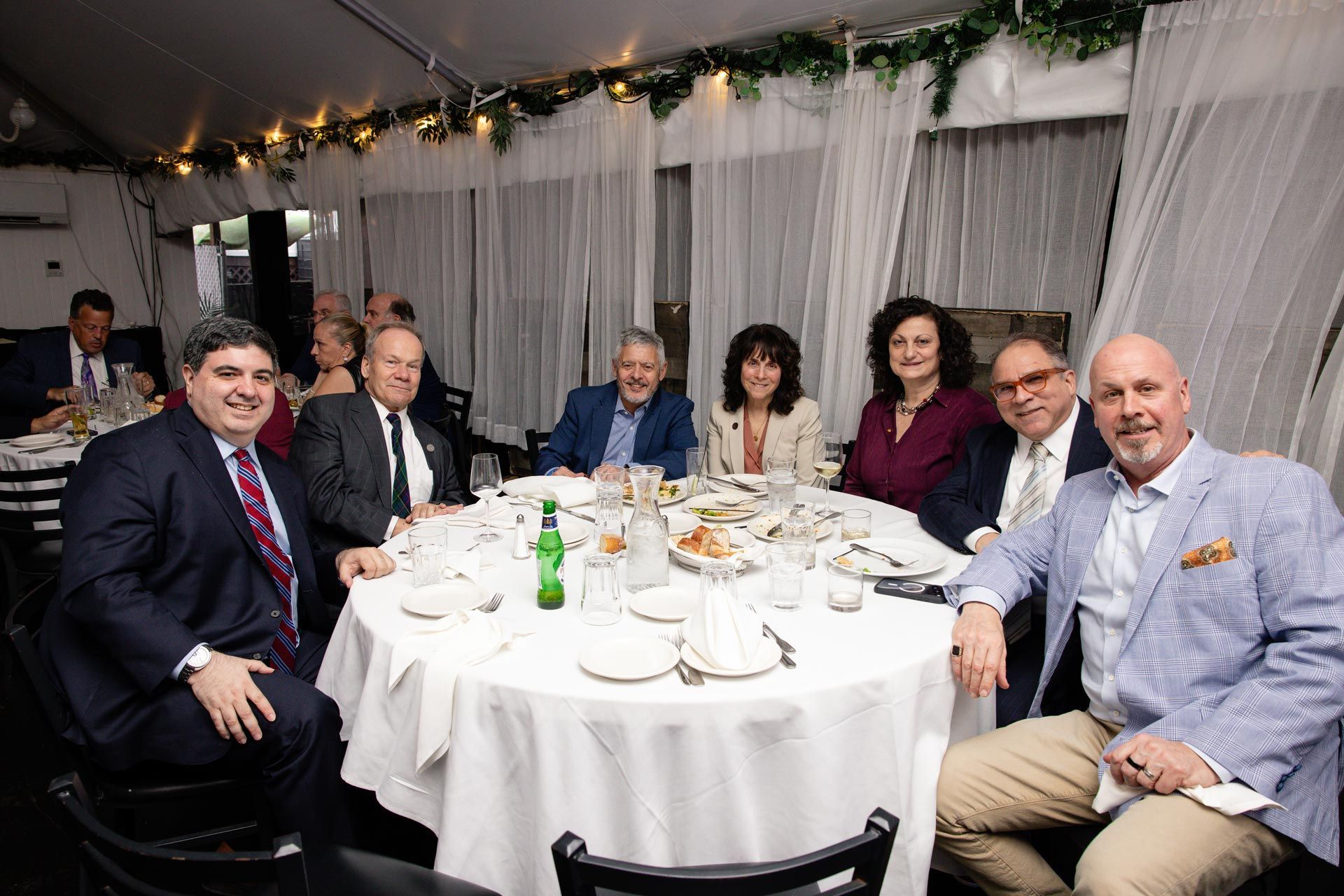 Seven people seated around a round banquet table at a formal event under string lights
