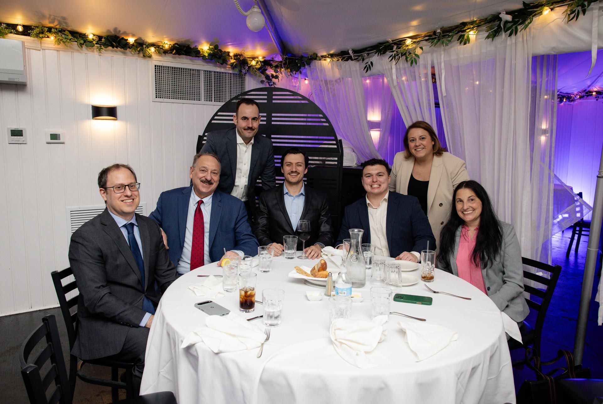 Seven people posing around a round table at a formal indoor event with string lights and white drapes.