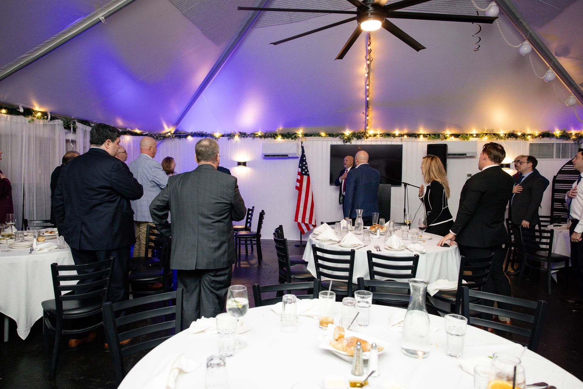 People standing at round tables inside a lit event tent with an American flag and stage area