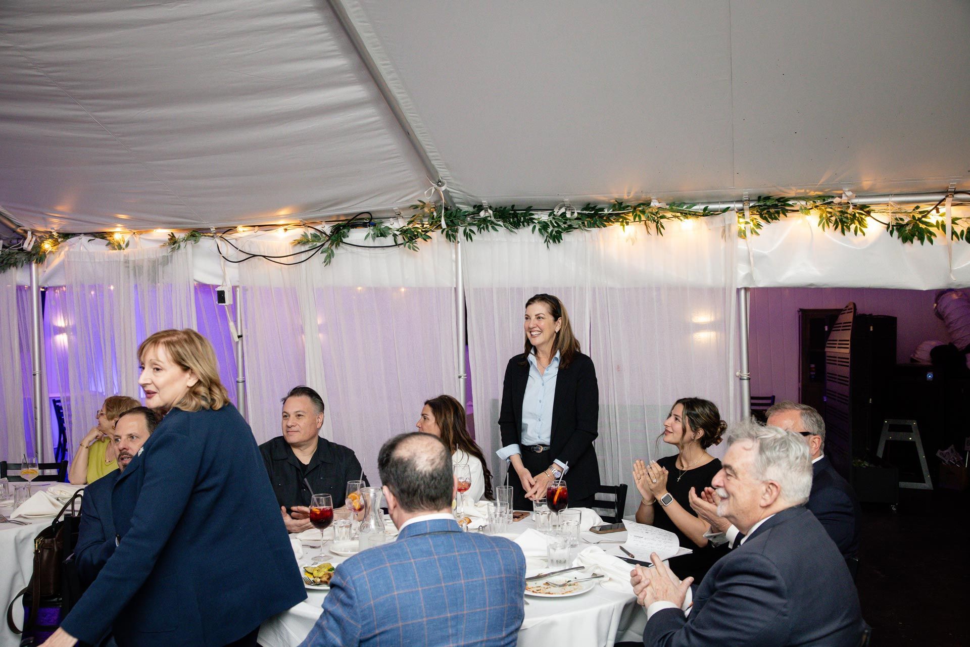 Guests seated at tables in a decorated tent, with a woman standing and speaking near the center.