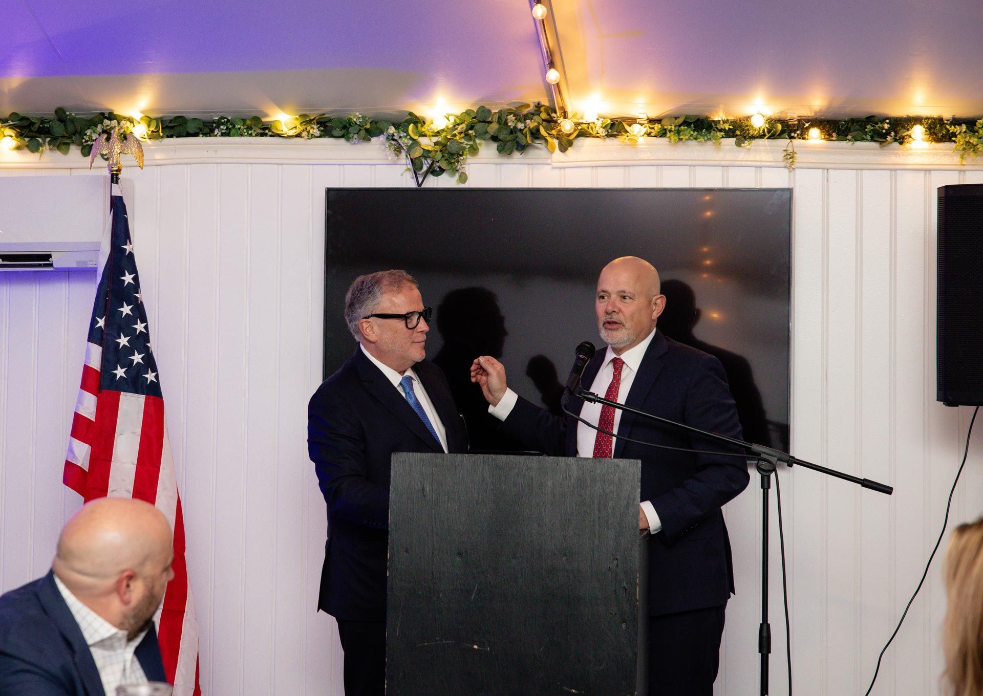 Two men at a podium in a decorated indoor venue, with an American flag and a microphone.