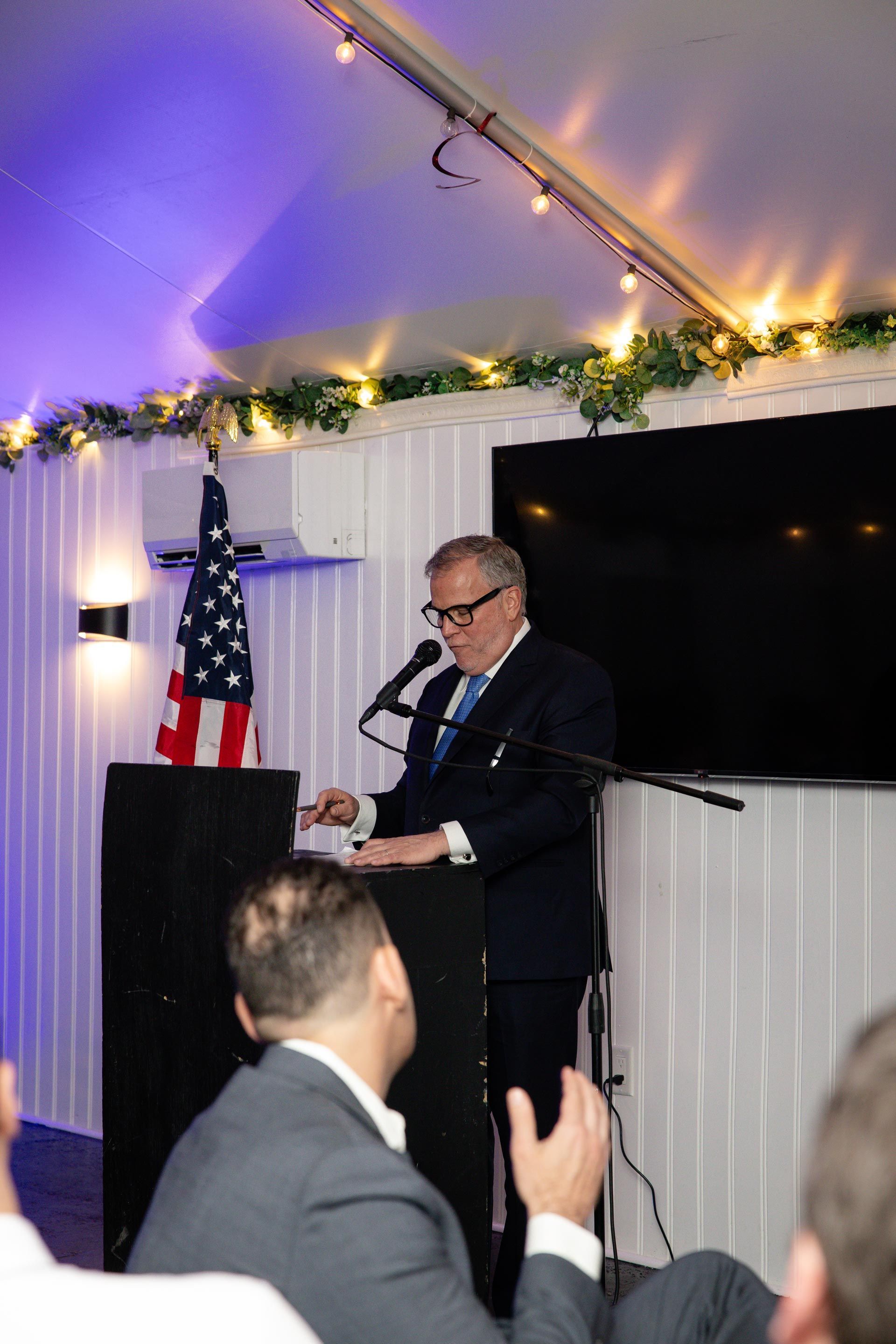 Man speaking at a podium under string lights at an indoor event, with an American flag nearby