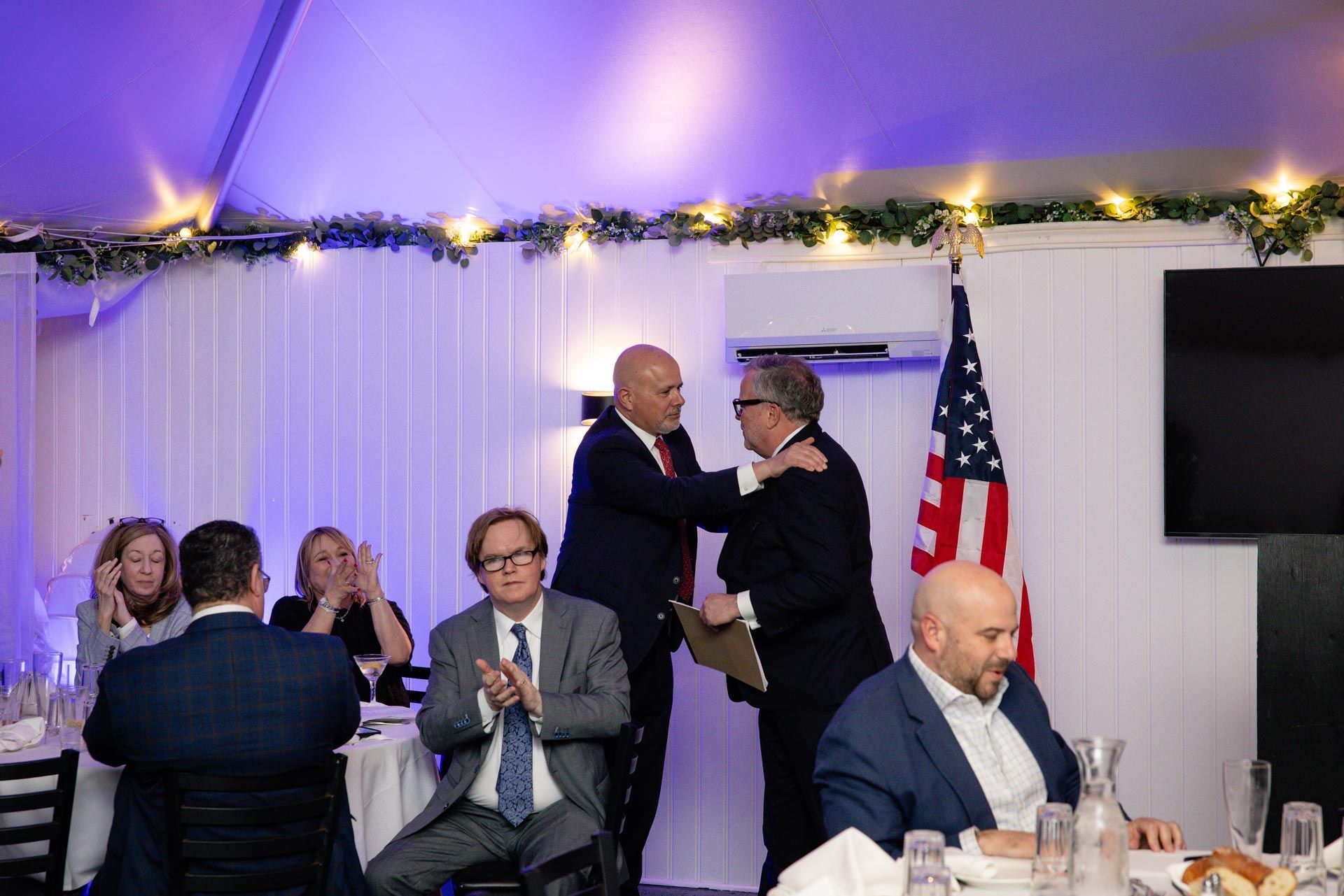 Two people embracing at a formal indoor event with seated guests and an American flag in the background