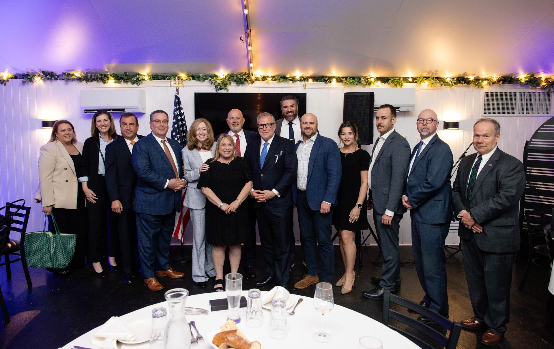 Group of people in formal wear posing at a banquet table under string lights at an indoor event