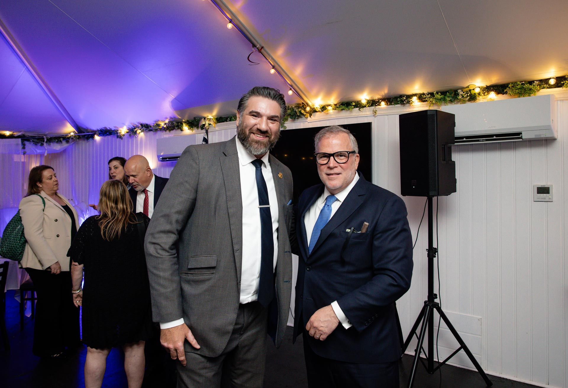 Two men in suits smiling at an indoor event under a tent with string lights.