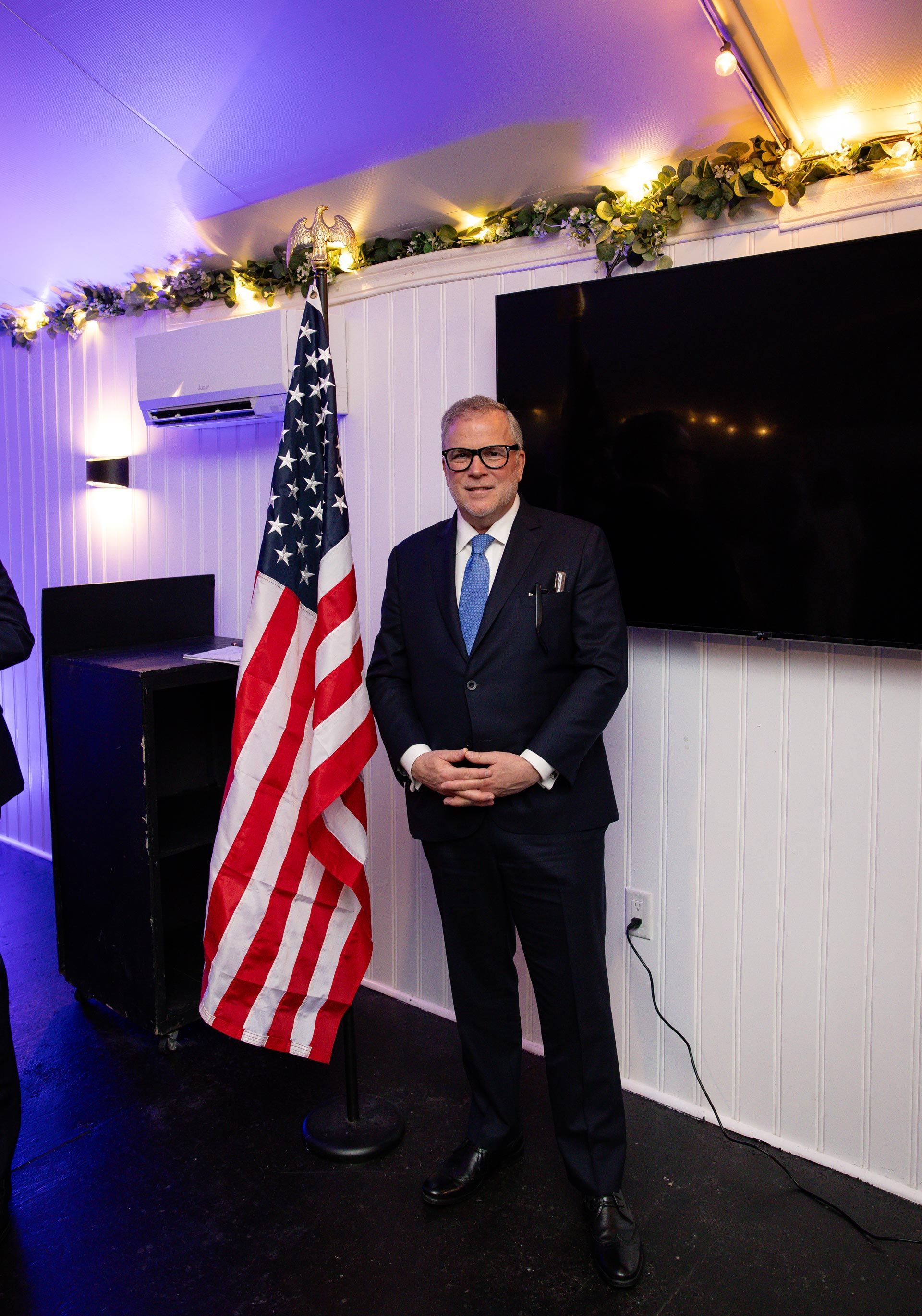 Man in suit stands beside an American flag on a small lit stage with garland overhead.