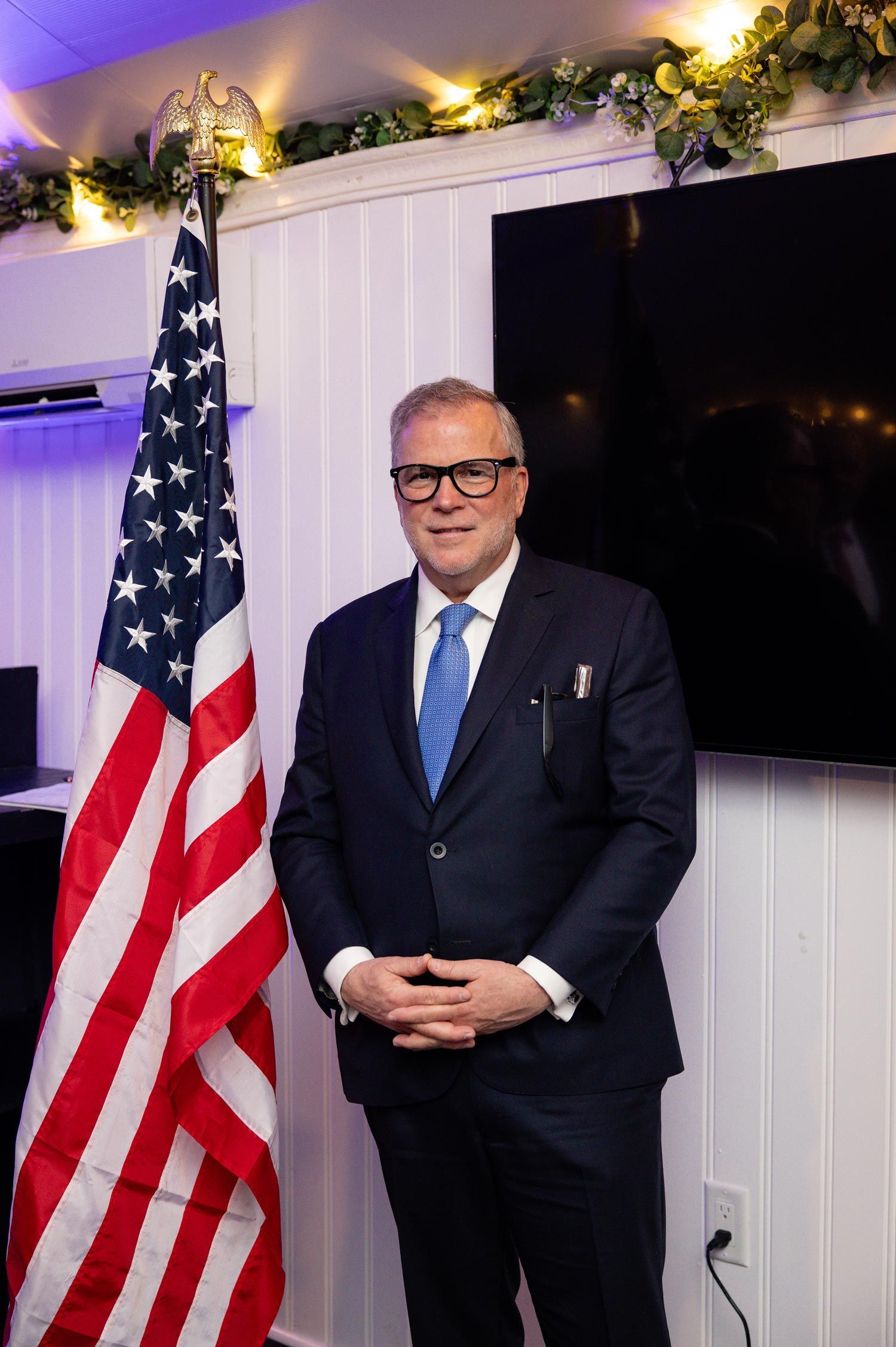 Man in a navy suit standing beside an American flag in a decorated indoor room