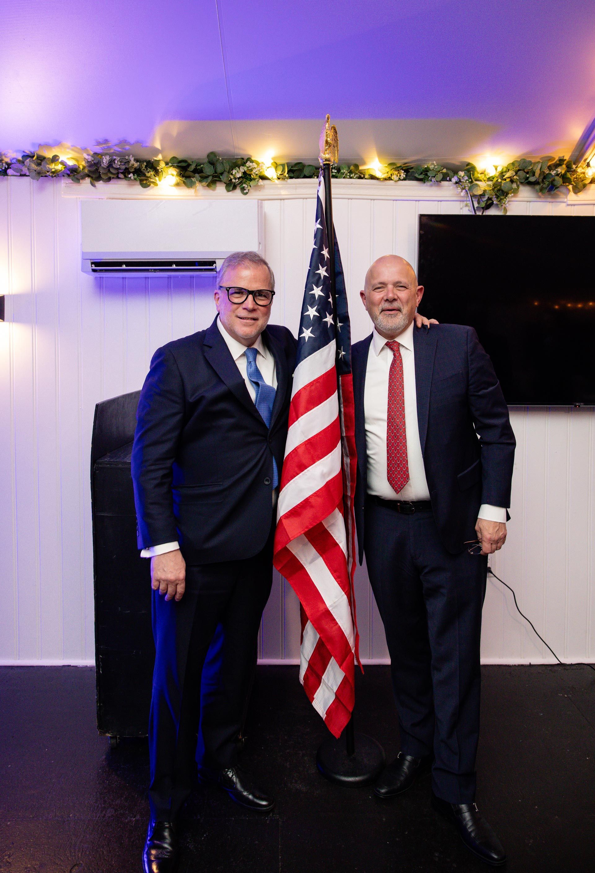 Two men in suits stand beside an American flag at an indoor event with string lights and a TV screen.